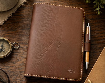 May include: A brown leather notebook cover with visible grain and light-colored stitching along the edges. A wooden pen is held in a side loop attached to the cover. A small embossed logo is visible on the bottom right corner of the front cover.