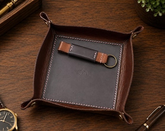 May include: A square dark brown leather valet tray with brass corner snaps and a central dark gray leather inlay featuring a small mountain logo stamp. A matching dark brown and black leather keychain with a brass ring rests inside the tray.