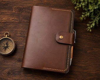 May include: A dark brown leather journal featuring a wrap-around strap with a brass snap button closure, visible tan stitching along the edges, and a pen holder containing a metallic pen, accompanied by a round brass compass with a black dial.