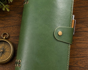 May include: A forest green leather journal featuring a snap button closure, visible stitching along the edges, and metallic eyelets on the spine. A wooden pen is tucked into the side, and the cover has a smooth, polished finish with a strap.