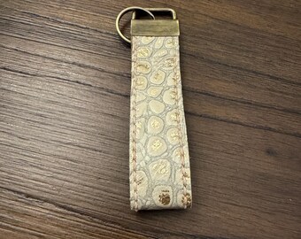 May include: A rectangular keychain strap featuring a textured off-white surface with a raised, embossed pattern and metallic gold accents. The strap is secured with a rectangular antique brass-toned metal clamp and a matching circular split ring.
