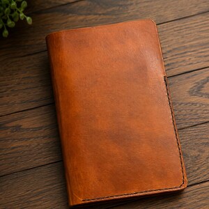 May include: A rectangular, tan-colored leather notebook cover with a smooth, slightly distressed finish. The cover features visible dark stitching along the edges, providing a clean, structured border for the folded leather construction.