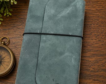 May include: A rectangular notebook with a matte, dusty blue-green cover featuring a wrap-around flap secured by a thin black elastic band. The notebook has a smooth, textured surface and is positioned vertically, showcasing its simple, functional design.