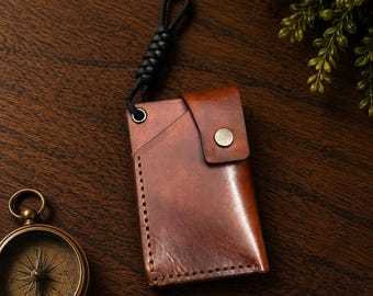 May include: A brown leather card holder featuring a snap button closure, visible stitching along the side, and a black braided cord lanyard attached to a metal eyelet. The leather has a smooth, polished finish with natural color variations.