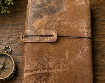 May include: A rectangular journal with a distressed brown leather cover featuring a textured surface, secured by a thin black elastic cord wrapped around a matching leather tab. A round brass compass with a black dial sits to the left.