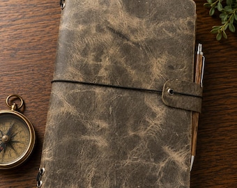 May include: A rectangular journal with a distressed, mottled brown leather cover featuring a black elastic band closure, a matching leather strap, and metal eyelets. A wooden pen is tucked into the side, and a brass compass sits nearby.