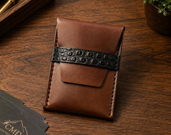 May include: A brown leather card holder featuring a smooth finish and a horizontal black strap with a textured crocodile-embossed pattern. The wallet is constructed with visible stitching along the sides and a flap closure design.