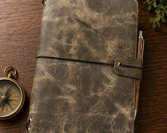 May include: A rectangular journal with a distressed, mottled brown leather cover featuring a black elastic band closure, a matching leather strap, and metal eyelets. A wooden pen is tucked into the side, and a brass compass sits nearby.