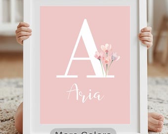 May include: A framed pink print featuring the name "Aria" in white script below a large white letter "A" and delicate floral accents. The words "More Colors" appear at the bottom. The print is held by a person.