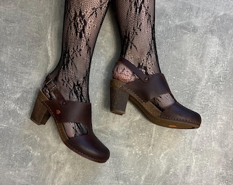 May include: Dark brown leather clogs with a wooden heel and ankle strap. The clogs feature a buckle fastening and a small flower detail. The shoes are worn with black lace stockings.