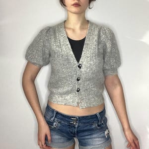 May include: A light gray, short-sleeved cardigan with a textured knit pattern and black buttons. The cardigan is paired with denim shorts. The top has a V-neck and a black undershirt is visible.