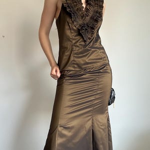 May include: A brown sleeveless formal gown with a ruffled neckline. The dress has a fitted bodice and a flared skirt with a slit. The fabric has a subtle sheen. A small black purse hangs on the side.