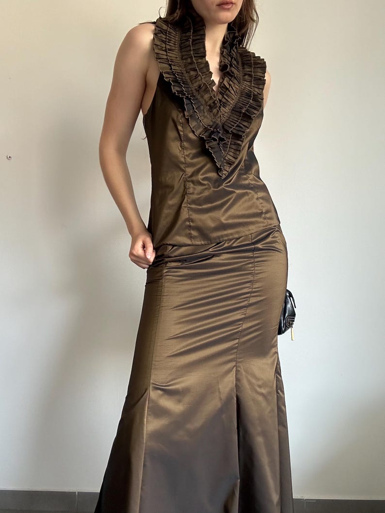 May include: A brown sleeveless formal gown with a ruffled neckline. The dress has a fitted bodice and a flared skirt with a slit. The fabric has a subtle sheen. A small black purse hangs on the side.