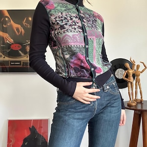 Può includere: Top a maniche lunghe con bottoni e motivo floreale e geometrico nei toni del rosa, verde e viola. Il top ha un collo alto ed è abbinato a jeans blu. Le maniche e il colletto sono neri.