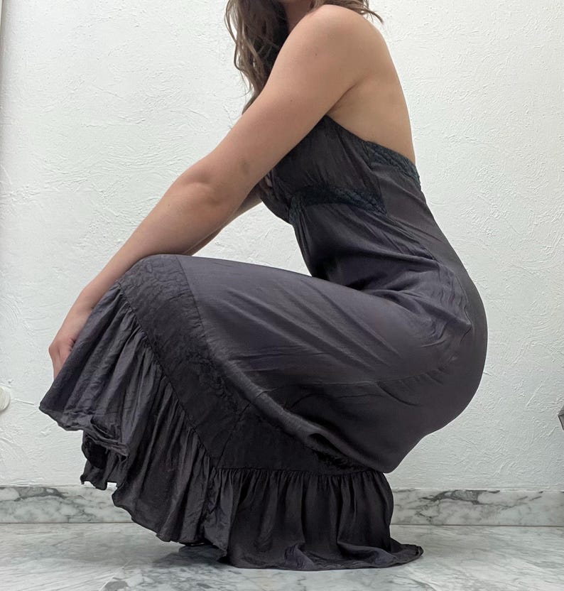 May include: A dark gray halter-neck dress with a flowing, ruffled hem. The dress features a fitted bodice with delicate lace detailing and a flared skirt. The fabric has a subtle sheen, creating a soft, elegant look. The dress is suitable for formal occasions.