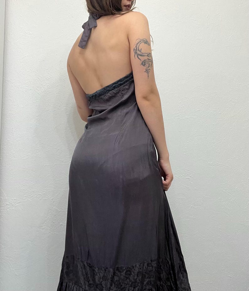 May include: A dark gray halter dress with a flowing silhouette. The dress features a low back and a decorative trim along the neckline. The hemline has a darker, textured pattern.