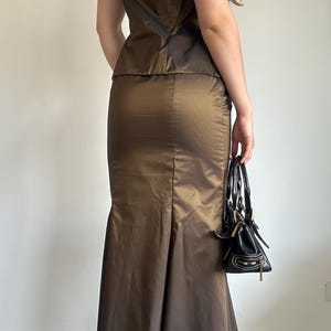 May include: A two-piece formal outfit in a bronze-colored fabric. The set includes a strapless top and a floor-length skirt with a flared hem. A small black handbag with white stitching is held in the hand.