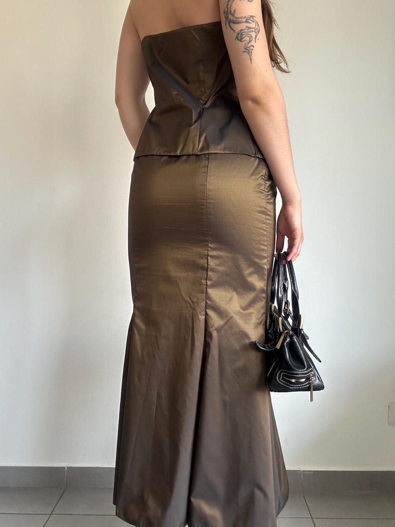 May include: A two-piece formal outfit in a bronze-colored fabric. The set includes a strapless top and a floor-length skirt with a flared hem. A small black handbag with white stitching is held in the hand.