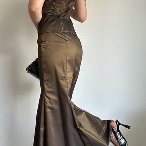 May include: A two-piece formal gown in a bronze-brown color. The strapless top and mermaid-style skirt are made of a shiny fabric. A black clutch and black high heels complete the ensemble. The dress is suitable for a special occasion.