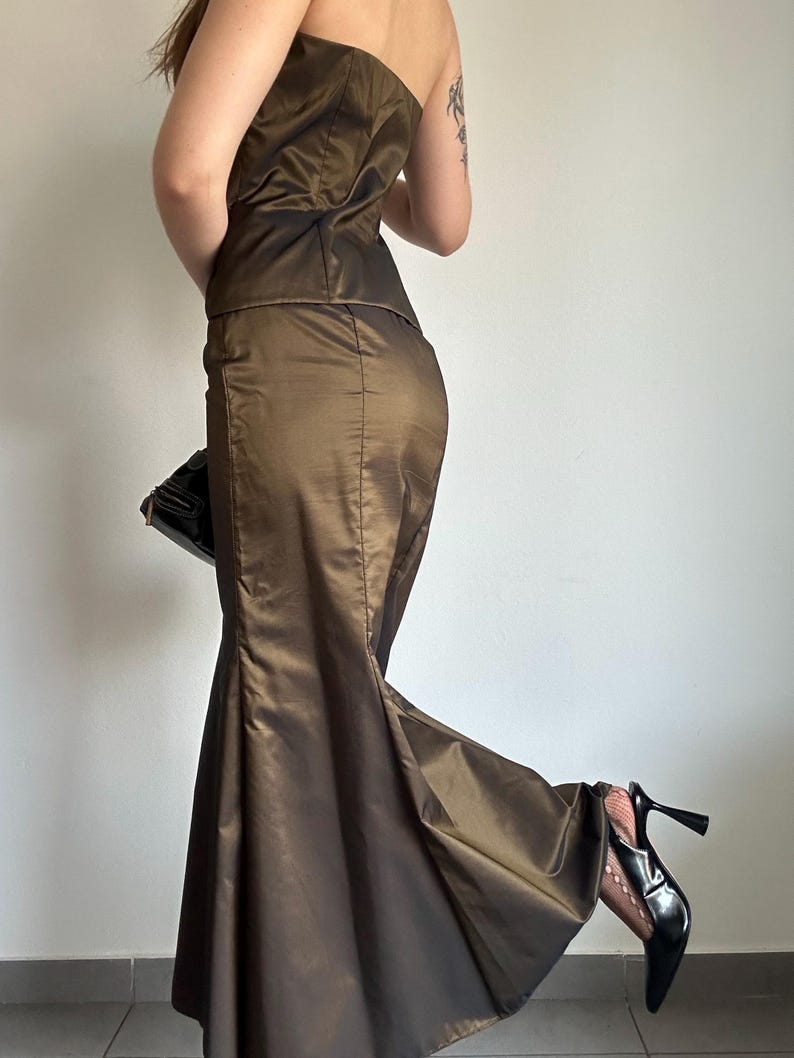 May include: A two-piece formal gown in a bronze-brown color. The strapless top and mermaid-style skirt are made of a shiny fabric. A black clutch and black high heels complete the ensemble. The dress is suitable for a special occasion.