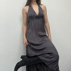 May include: A dark gray halter maxi dress with a V-neckline and a flowing skirt. The dress has a fitted bodice with a decorative trim and a tiered hem. The model is wearing black boots.