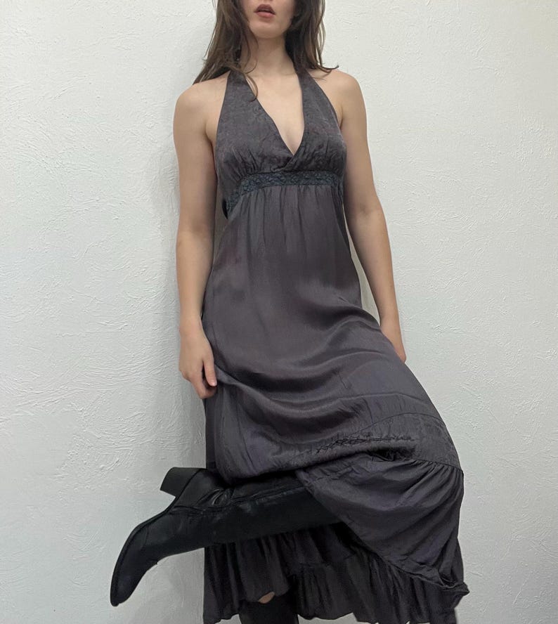 May include: A dark gray halter maxi dress with a V-neckline and a flowing skirt. The dress has a fitted bodice with a decorative trim and a tiered hem. The model is wearing black boots.