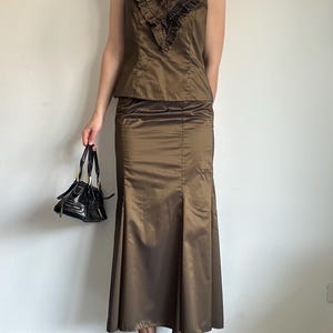 May include: A two-piece ensemble featuring a sleeveless top with a ruffled neckline and a matching floor-length skirt. The outfit is a metallic bronze color. A small, black handbag with gold hardware is held in the hand.
