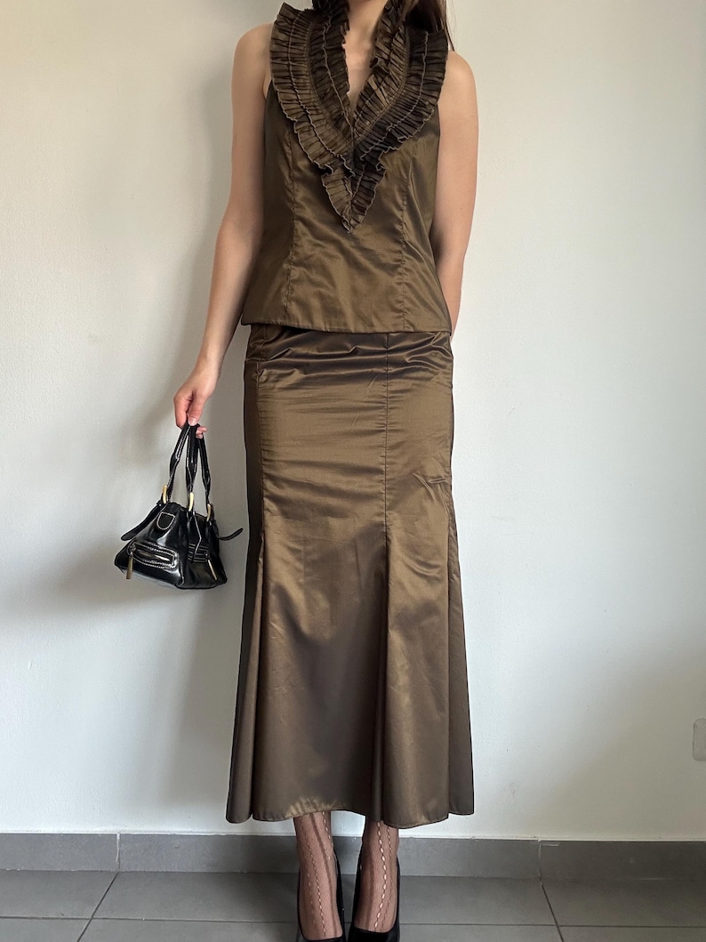 May include: A two-piece ensemble featuring a sleeveless top with a ruffled neckline and a matching floor-length skirt. The outfit is a metallic bronze color. A small, black handbag with gold hardware is held in the hand.