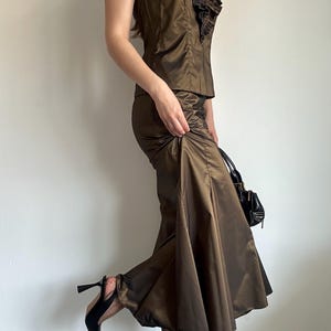 May include: A brown satin two-piece outfit featuring a halter top with ruffled detailing and a matching skirt. The outfit is paired with black heels and a small black handbag. The fabric has a subtle sheen, suggesting a formal or evening wear style.