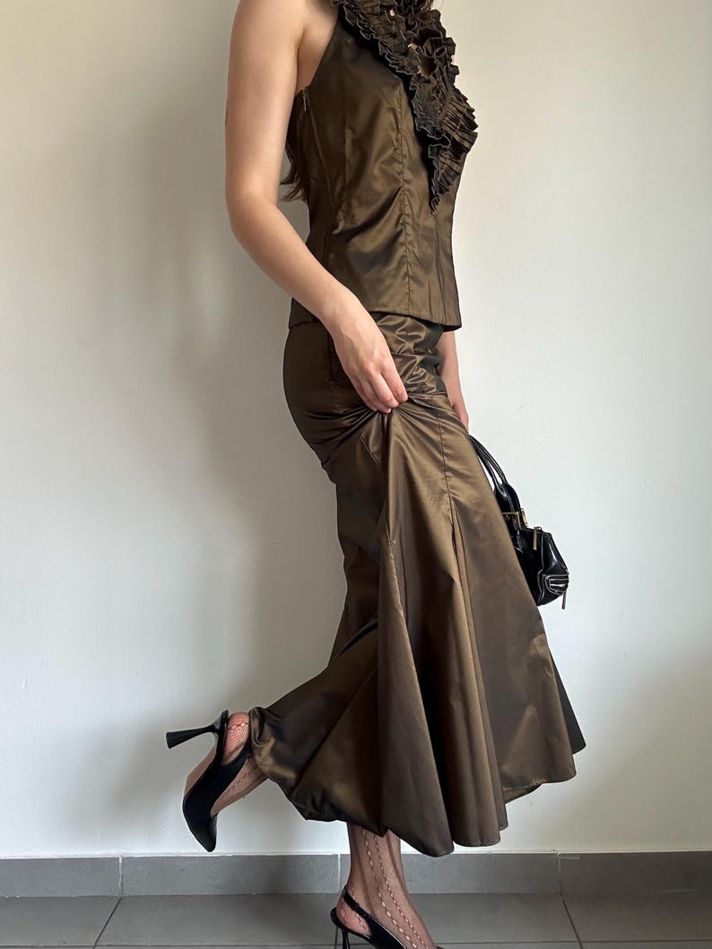 May include: A brown satin two-piece outfit featuring a halter top with ruffled detailing and a matching skirt. The outfit is paired with black heels and a small black handbag. The fabric has a subtle sheen, suggesting a formal or evening wear style.