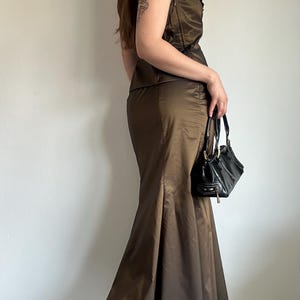 May include: A brown satin two-piece outfit, featuring a sleeveless top with ruffled trim and a floor-length, flared skirt. A black handbag with a top handle is held. The outfit is paired with black high-heeled shoes.