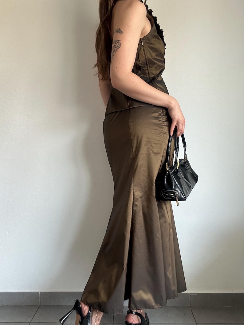 May include: A brown satin two-piece outfit, featuring a sleeveless top with ruffled trim and a floor-length, flared skirt. A black handbag with a top handle is held. The outfit is paired with black high-heeled shoes.