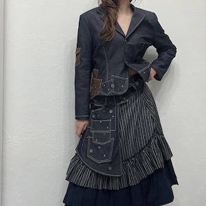 Puede incluir: Chaqueta vaquera azul oscuro con parches marrones y falda a juego. La falda presenta un panel a rayas y un dobladillo con volantes. El conjunto es de color azul oscuro.