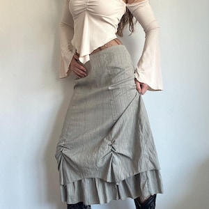 May include: A beige off-the-shoulder top with long sleeves and a gathered bodice. The outfit includes a light green skirt with vertical stripes, ruched detailing, and a ruffled hem. The skirt is paired with black boots.