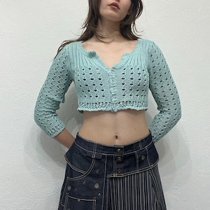Könnte beinhalten: Hellblaues, kurzes, langärmliges Strickjäckchen mit V-Ausschnitt und Knopfverschluss. Das Strickjäckchen hat ein zartes Lochmuster. Das Model trägt einen Jeansrock mit Streifenmuster.
