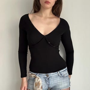 May include: A black long-sleeve top with a deep V-neckline and button details. The top is form-fitting and has a fitted silhouette. The model is wearing denim shorts. The top is made of a soft, stretchy material.