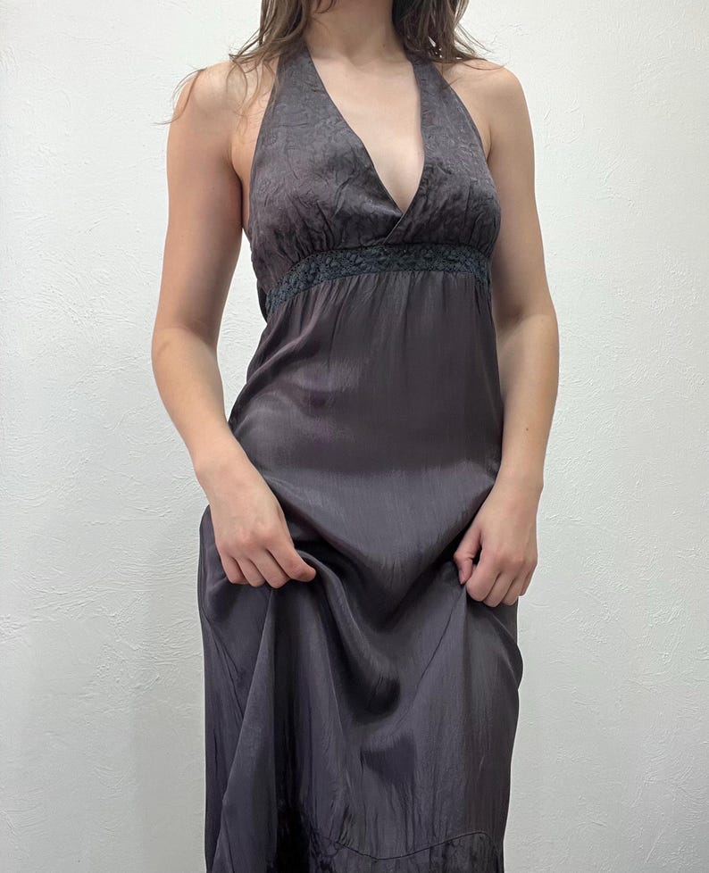 May include: A dark grey halter dress with a deep V-neckline. The dress features a textured bodice and a flowing skirt. A dark lace trim accents the waistline. The dress is made of a silky material and has a subtle sheen.