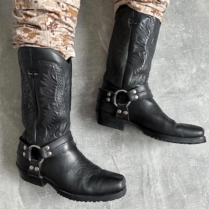 Op de afbeelding: Een paar zwarte leren harnaslaarzen met gedetailleerde ton-sur-ton stiksels op de schacht, een zilverkleurig metalen ringdetail met klinknagels, een ronde neus en een lage blokhak, gedragen met een broek met camouflagepatroon.