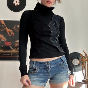 Puede incluir: Camiseta negra de manga larga con cuello alto y chaleco vaquero. El chaleco tiene un diseño desgastado con remaches plateados. Se usa con shorts de mezclilla con detalles de pedrería. El atuendo tiene una estética punk rock.