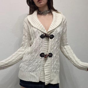 Peut inclure: Cardigan en tricot de câble crème avec un col châle et des fermetures à bascule. Le pull à manches longues présente un motif texturé et une coupe décontractée, adapté au superposition. Les fermetures à bascule sont marron foncé.