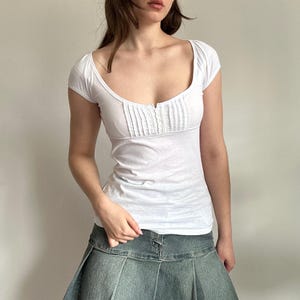 May include: White short-sleeve top with a scoop neckline and pleated detailing on the bodice. The top has a fitted silhouette and a button closure. The fabric appears to be a lightweight material, suitable for warmer weather. The top is paired with a denim skirt.