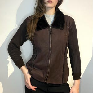 Puede incluir: Chaqueta marrón con cuello de piel sintética negra y cremallera completa. La chaqueta tiene mangas de punto acanalado y un cuerpo similar al ante. Pequeños tachuelas plateadas son visibles en la parte delantera.
