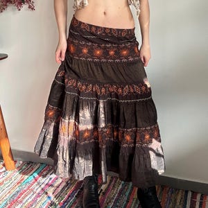 May include: A long, tiered brown skirt with embroidered floral and starburst designs in orange. The skirt has a flowing, bohemian style, with multiple layers and a wide waistband. The skirt is made of a soft, flowing fabric.
