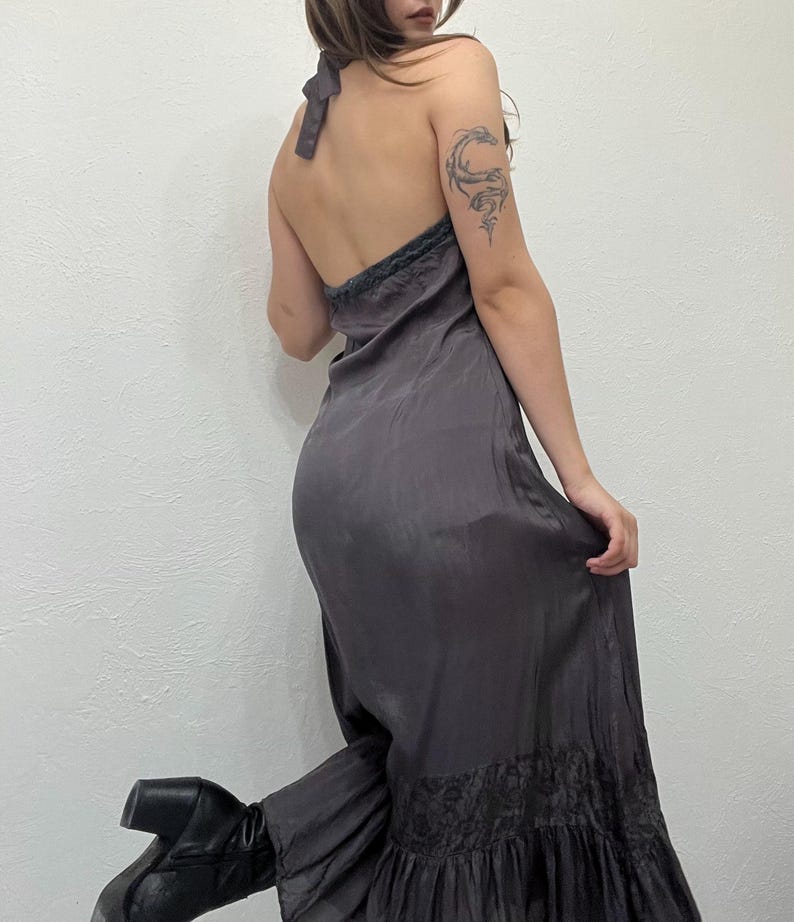 May include: A dark gray halter dress with a flowing silhouette. The dress features a delicate lace trim along the neckline and hem. The dress is made of a silky material and has a subtle sheen. The dress is paired with black boots.