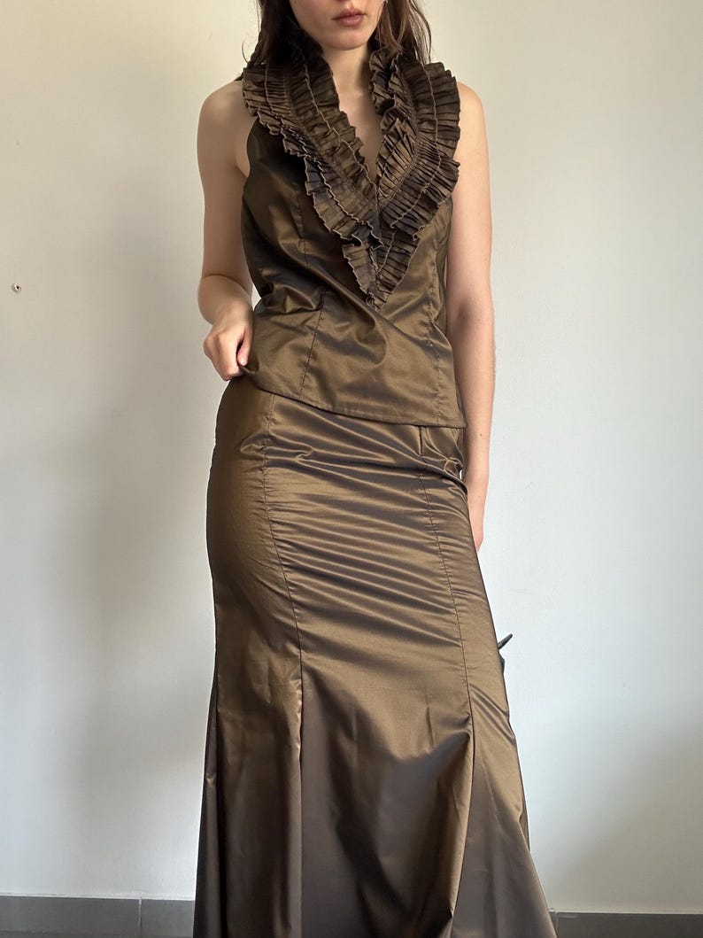 May include: A two-piece formal outfit in a bronze color. The top is a halter-style with a ruffled collar. The skirt is a floor-length, fitted design with a slight flare at the bottom. The fabric has a subtle sheen.