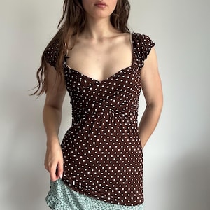 May include: A brown top with a white polka dot pattern, featuring short cap sleeves, a sweetheart neckline, and a gathered crossover bodice design that extends down to the waistline, creating a fitted silhouette with a flared hemline.