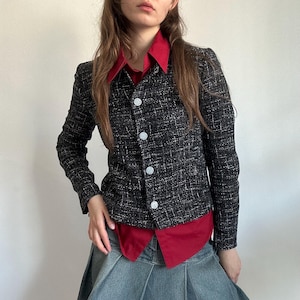 May include: A black and white tweed jacket with three white buttons. The jacket is worn over a red collared shirt and paired with a denim skirt. The jacket has a textured appearance.