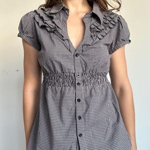 May include: A black and white gingham button-down shirt featuring short puff sleeves, a collared neckline, and layered ruffle detailing along the front placket. The garment includes a smocked elasticated waistline and a central button closure.