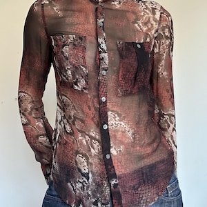 May include: A long-sleeved, button-down shirt made of a sheer, semi-transparent fabric featuring a dark red, black, and beige snakeskin print. The shirt includes a mandarin collar, a front button placket, and two chest pockets with button flaps.