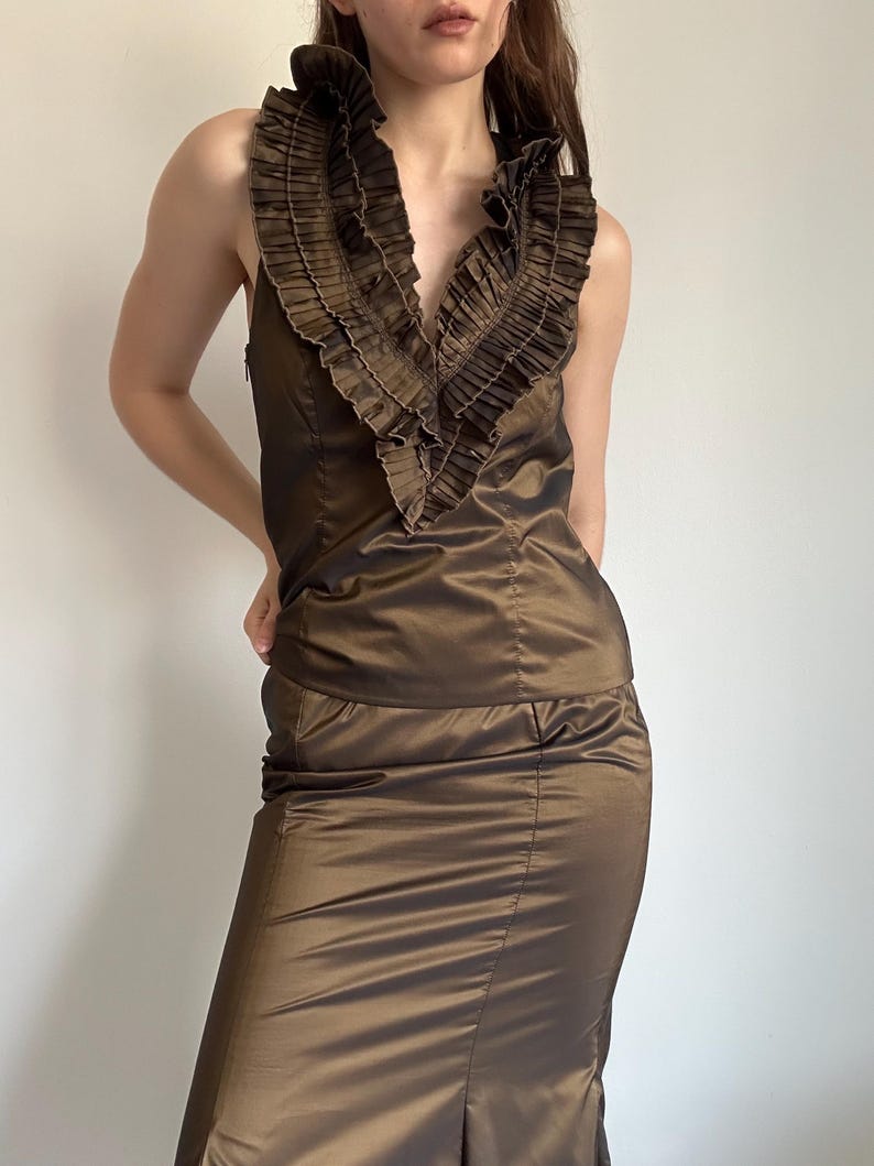 May include: A brown satin two-piece outfit. The sleeveless top features a deep V-neckline with a ruffled collar. The matching skirt is knee-length with a slight slit in the back. The outfit has a sheen and a tailored fit.